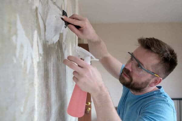 Home repairs. Focused man removes the old wallpaper from the wall with a spatula and a pulverizer with water.